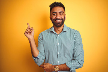 Young indian man wearing green striped shirt standing over isolated yellow background with a big smile on face, pointing with hand and finger to the side looking at the camera.