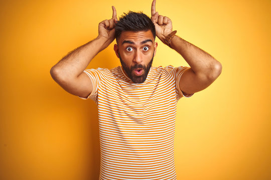Young indian man wearing t-shirt standing over isolated yellow background doing funny gesture with finger over head as bull horns