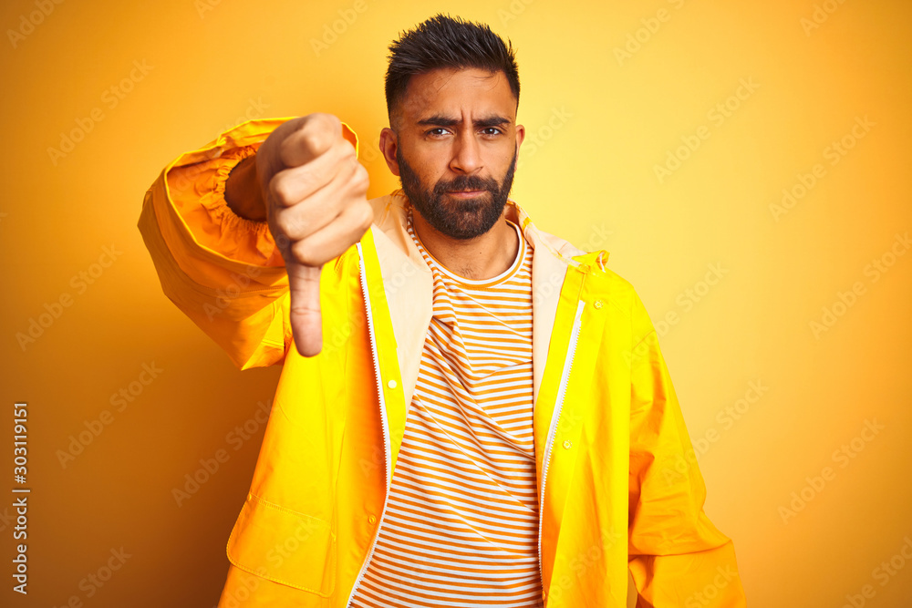 Sticker Young indian man wearing raincoat standing over isolated yellow background looking unhappy and angry showing rejection and negative with thumbs down gesture. Bad expression.