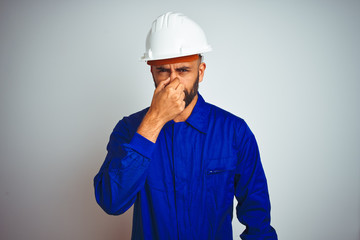 Handsome indian worker man wearing uniform and helmet over isolated white background smelling something stinky and disgusting, intolerable smell, holding breath with fingers on nose. Bad smells 
