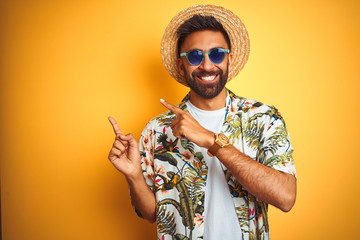 Indian man on vacation wearing floral shirt hat sunglasses over isolated yellow background smiling...