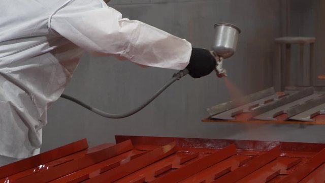 Jet of red paint is sprayed from a gun, worker paints metal in a workshop, spraying, protecting parts, close-up, protective suit and gloves, work is in full swing, professional spray gun, airbrushing