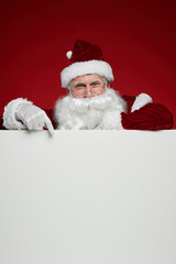 Portrait of Santa Claus with white beard looking at camera and pointing at blank billboard isolated on red background