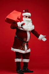 Fototapeta na wymiar Portrait of Santa Claus in sunglasses holding red sack with Christmas presents and standing against the red background