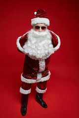 Portrait of Santa Claus in red costume and with white beard posing at camera in sunglasses against the red background