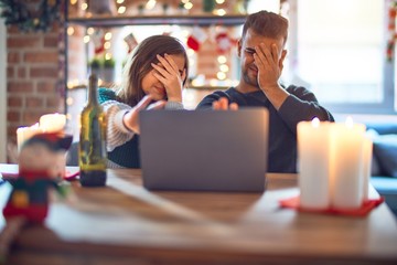 Young beautiful couple sitting using laptop around christmas decoration at home covering eyes with hands and doing stop gesture with sad and fear expression. Embarrassed and negative concept.
