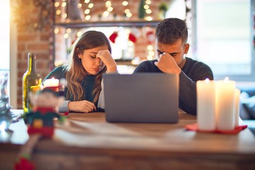 Young beautiful couple sitting using laptop around christmas decoration at home tired rubbing nose...
