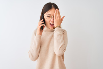 Young chinese woman talking on the smartphone over isolated white background covering one eye with hand, confident smile on face and surprise emotion.
