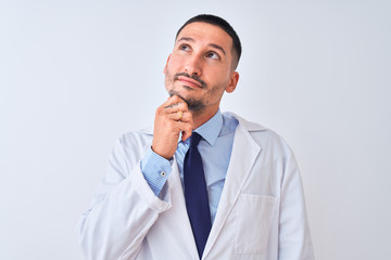 Young doctor man wearing white coat over isolated background with hand on chin thinking about question, pensive expression. Smiling with thoughtful face. Doubt concept.