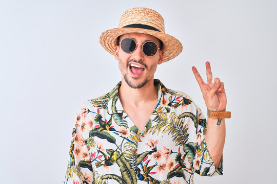 Young handsome man wearing Hawaiian shirt and summer hat over isolated background smiling with happy face winking at the camera doing victory sign. Number two.