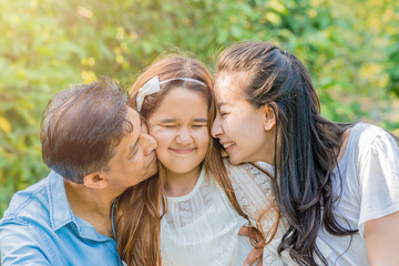 Fototapeta premium Asian parents kissing their daughter in the cheeks outdoor.