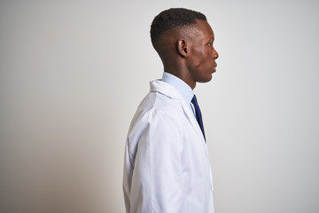 Young african american doctor man wearing coat standing over isolated white background looking to side, relax profile pose with natural face with confident smile.