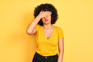 Fototapeta na wymiar Young arab woman with curly hair wearing t-shirt standing over isolated yellow background covering eyes with hand, looking serious and sad. Sightless, hiding and rejection concept
