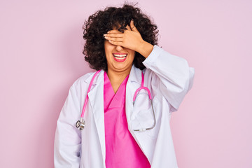 Young arab doctor woman with curly hair wearing stethoscope over isolated pink background smiling and laughing with hand on face covering eyes for surprise. Blind concept.