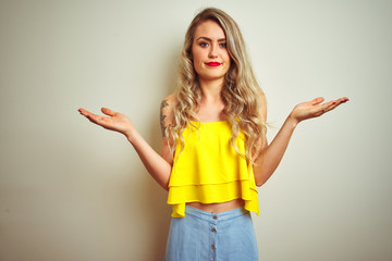 Young beautiful woman wearing yellow t-shirt standing over white isolated background clueless and confused expression with arms and hands raised. Doubt concept.