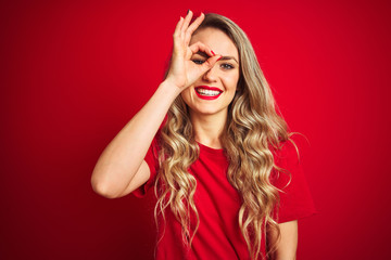 Young beautiful woman wearing basic t-shirt standing over red isolated background doing ok gesture with hand smiling, eye looking through fingers with happy face.