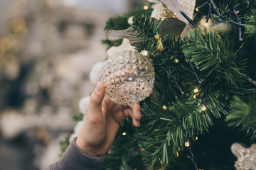 Young boy looking Christmas tree decorations, thinking about Santa's gifts. Kid decorating with baubles a branch of spruce tree on bright blurred background. Holidays, party and new year eve concept.