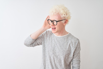 Young albino blond man wearing striped t-shirt and glasses over isolated white background smiling with hand over ear listening an hearing to rumor or gossip. Deafness concept.