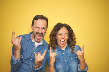 Beautiful middle age couple together standing over isolated yellow background shouting with crazy expression doing rock symbol with hands up. Music star. Heavy music concept.