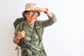 Fototapeta premium Middle age hiker woman wearing backpack canteen hat glasses over isolated white background very happy and smiling looking far away with hand over head. Searching concept.