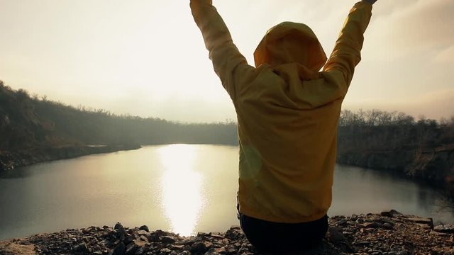 Traveler woman looking at sunset sitting near lake with raised hands enjoying freedom