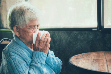 Old person coughing covering mouth with a tissue on a house interior. She has flu, allergy symptoms, acute bronchitis, pulmonary infections or pneumonia. Concept:respiratory infections and healthcare.