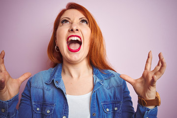 Youg beautiful redhead woman wearing denim shirt standing over isolated pink background crazy and mad shouting and yelling with aggressive expression and arms raised. Frustration concept.