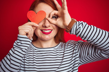 Young redhead romantic woman holding heart over red isolated background with happy face smiling doing ok sign with hand on eye looking through fingers
