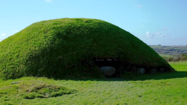 Handheld wide shot of one of the Tombs of Knowth covered with green grass against a blue sky on a sunny day. Isolated shot of the exterior tomb with no people.