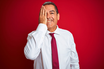 Handsome middle age businessman standing over isolated red background covering one eye with hand, confident smile on face and surprise emotion.