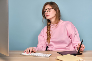 A girl in pink sweatshirt with a braid and sits at a table and looks at the monitor, holding a...