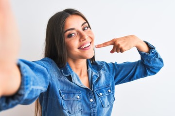 Beautiful woman wearing denim shirt make selfie by camera over isolated white background smiling cheerful showing and pointing with fingers teeth and mouth. Dental health concept.