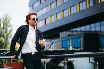 A Man With A Nice Beard Is Standing In The Street In A Blue Suit And An Orange Leather Bag While Drinking His Coffee