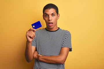 Young handsome arab man holding credit card standing over isolated yellow background scared in shock with a surprise face, afraid and excited with fear expression