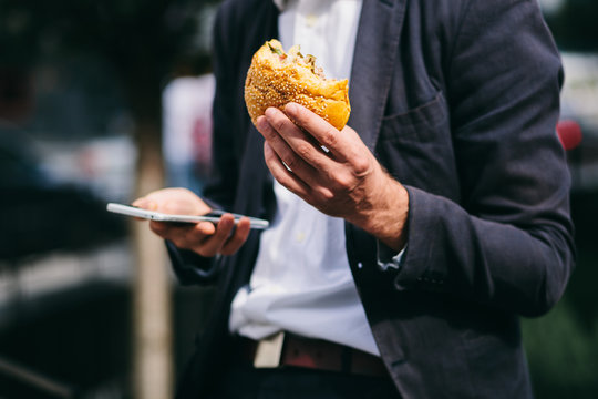 Hamburger And Mobile Phone In The Hand Of A Businessman Close-up Shot.