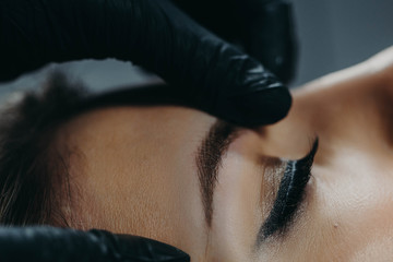 A microblading master in black rubber gloves holds a close-up of a girl’s client’s finished...