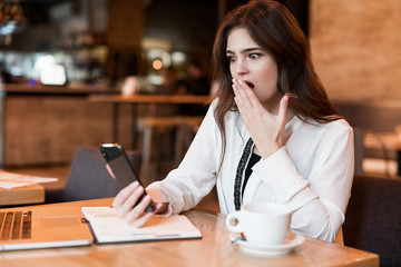 young beautiful woman with phone in her hand working outside office in laptop looks shocked recieving bad news drinking hot coffee in cafe modern businesswoman