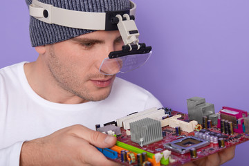Image of Caucasian young technician using magnifying glass for repairing personal computer's motherboard in his lab. Concept of computer hardware, mobile phone, electronic, repairing and technology.