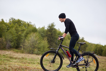 Fototapeta na wymiar Image of fit male mountain biker riding his bike uphill on meadow near forest, sporty guy wearing black track suit, spending time in open air, having active recreation. Healthy lifestyle concept.