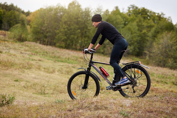 Fototapeta na wymiar Outdoor shotof man riding mountain bike in meadow, sporty male dresses black track suit and cap, spending free time in active way, enjoying cycling, young sportsman covering assigned distance.