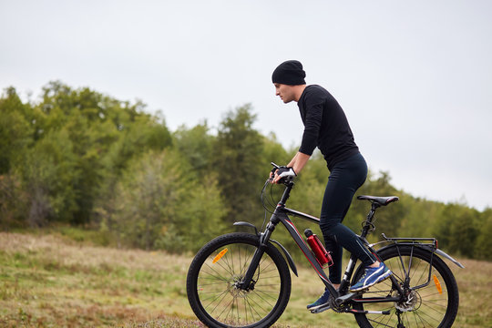 Image of fit male mountain biker riding his bike uphill on meadow near forest, sporty guy wearing black track suit, spending time in open air, having active recreation. Healthy lifestyle concept.