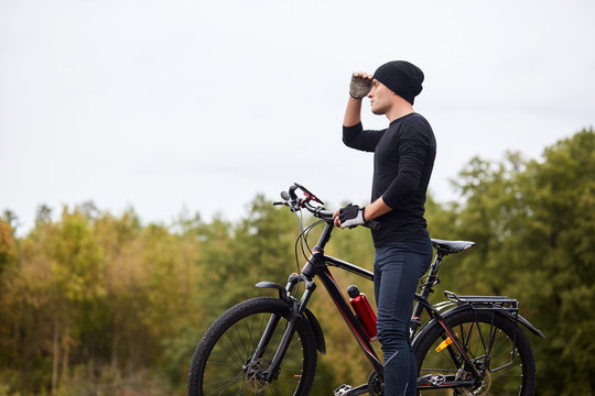 Image of road biker cycling and training on road in forest. Attractive young sportsman stops to have rest after long hours riding, keeps hand near forehead and looks faraway. Healthy activity concept.