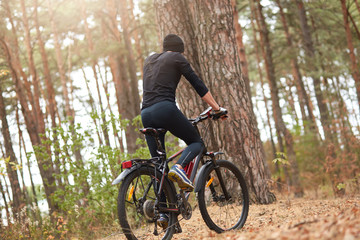 Fototapeta na wymiar Back view of biker on trail in woods, young man cycling MTB in forest, handsome man wearing black track suit spending time in open air, male riding uphill. Sport, fitness, motivation concept.