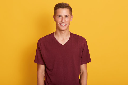 Close up portrait of pleased young guy dressed maroon casual t shirt, looking smiling at camera, expresses happyness, model posing isolated over yellow background. People, youth and lifestyle concept.