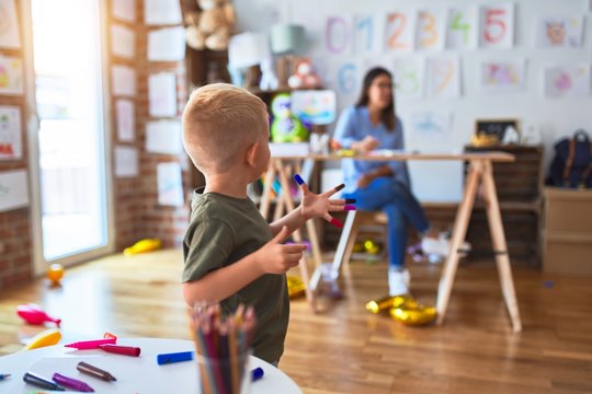 Young caucasian child playing at playschool with teacher. Mother and son at playroom drawing a draw with color pencils, young woman at the background sitting on desk.