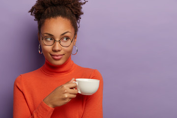 Charming dark skinned woman with curly crisp hair, drinks coffee or tea, holds white mug, wears spectacles and orange turtleneck, looks aside, isolated over purple background, free space right