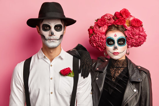 Horizontal shot of spooky couple dressed for Day of Dead in Mexico, wear skull masks, stand next to each other, celebrate Halloween together, isolated over pink background, have image of zombie