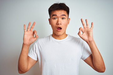 Young asian chinese man wearing t-shirt standing over isolated white background looking surprised and shocked doing ok approval symbol with fingers. Crazy expression