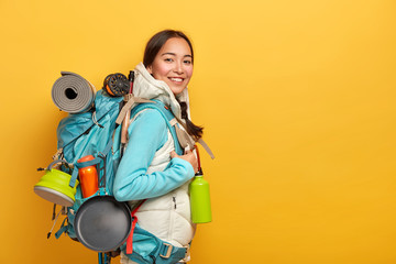 Positive Asian female hiker stands sideways to camera, carries big rucksack with necessary things for travel, has exciting adventurous trip, isolated over yellow wall, blank space on right side.