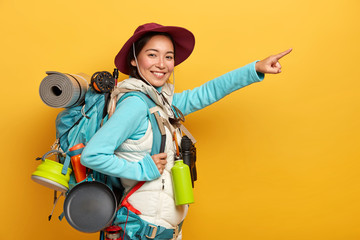 Glad smiling hiker dressed casually, stands with backpack against yellow background, points into distance and shows way to people, looks for direction while traveling, wears binoculars on neck